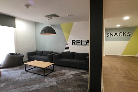 A room with the word 'relax' painted on a wall, two sofas, a coffee table and a bean bag. A wall in the corridor outside the room has the word 'snacks' painted on it.