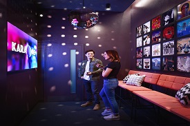 A man and a woman stood together smiling in a room with a mirrorball on the ceiling, seating behind them and many classic albums framed on the wall, and a screen with the word 'Karaoke' on it in front of them.