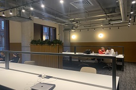 Study area with a series of long tables and a few students sitting at them. There is lighting and plug sockets.
