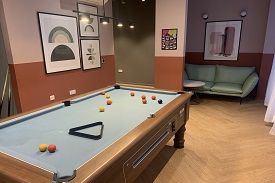 Pool table in the front left and a sofa in the background. Pictures on the wall and there is a light hanging down above the pool table.