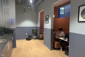 To the left is a vending machine. To the right are alcoves with tables and seats for people to meet in. One person is sitting in the middle alcove studying.