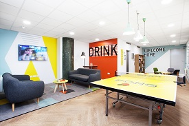 A room with a television screen on a wall, two sofas and a ping pong table. Ther word 'drink' is written on another wall. In the background there is another wall with 'welcome home' written on it, a bookcase, and three armchairs around a coffe table.