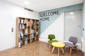 A room with a large bookcase, two armchairs and a coffee table. The words 'welcome home' are written on one wall.