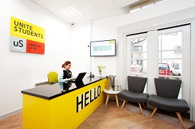 A woman using a laptop and sitting at a reception desk with the word 'hello' written on it. There are two armchairs nearby and the words 'Unite Students Chantry Court' on a sign behind her.
