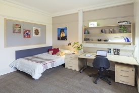 A room with a double bed in one corner and a desk next to it. The desk has an office chair beside it, drawers beneath it and shelves above it.