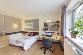 A room with a double bed in one corner and a desk with an office chair in another corner. The desk has two shelves above it and the bed has a chest of drawers next to it.