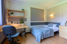 A room with a single bed in one corner and a desk and office chair opposite. There are shelves above the desk.
