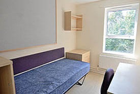 A room with a single bed against one wall and a desk and office chair opposite. There is a small chest of drawers at the foot of the bed and a set of shelves on the wall above it.