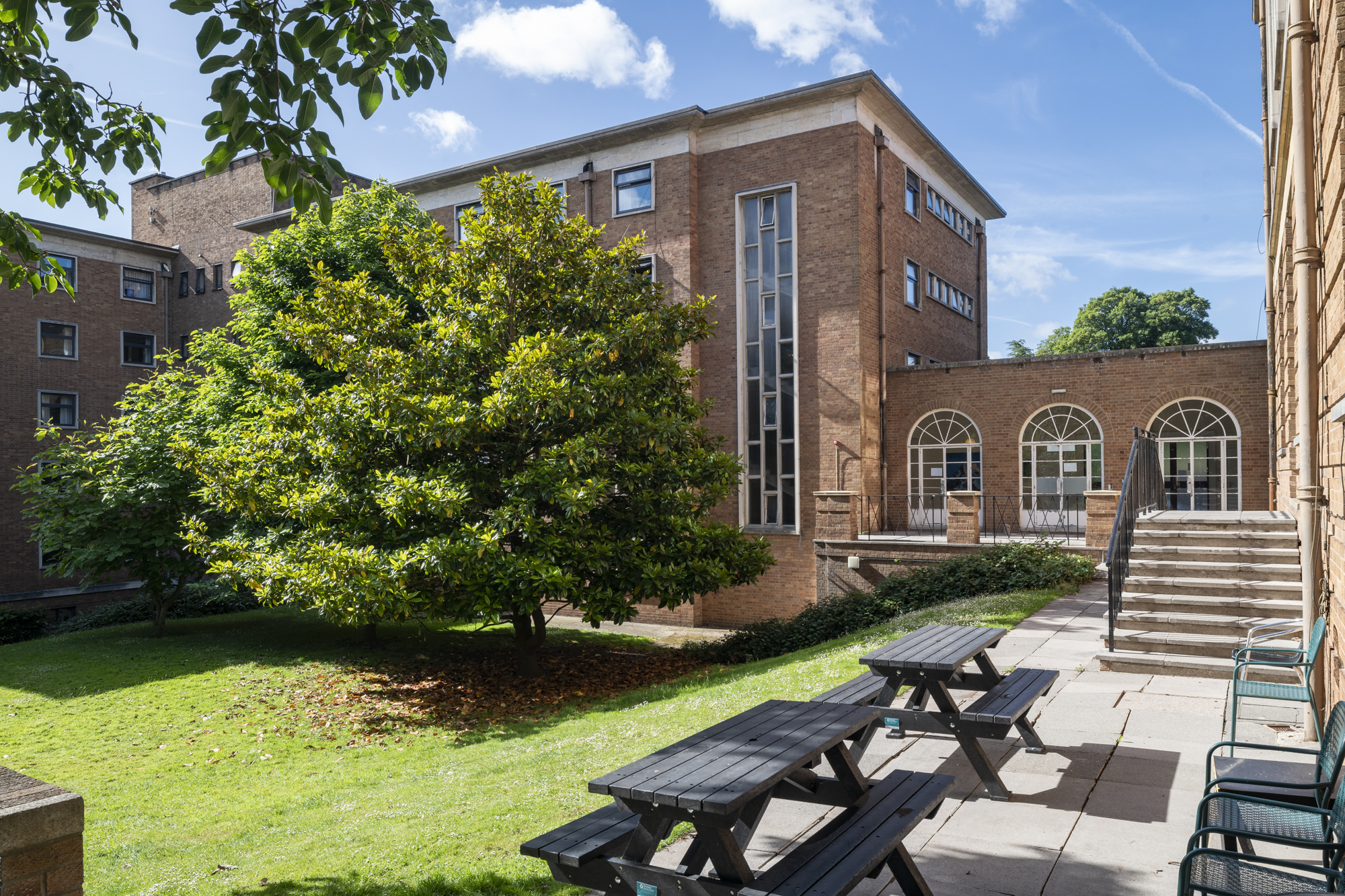 A lawn tree in front of a red brick building. A patio with picnic tables, and steps.