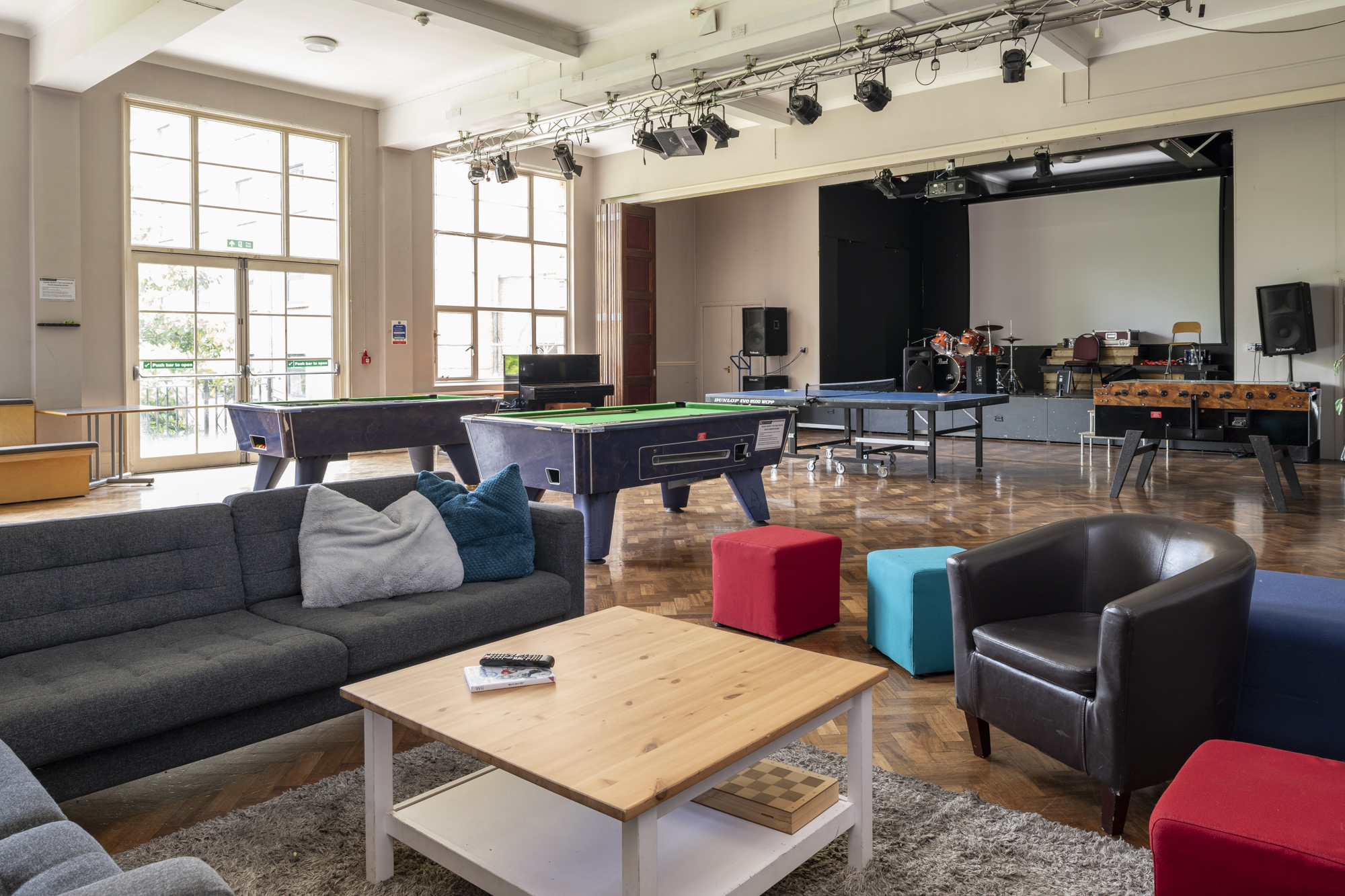 A large open room with large windows. In the foreground are sofas an arm chairs surrounding a coffee table. In the middleground behind are two snooker tables, a table tennis table, and table football. At the very back is a stage with instruments.