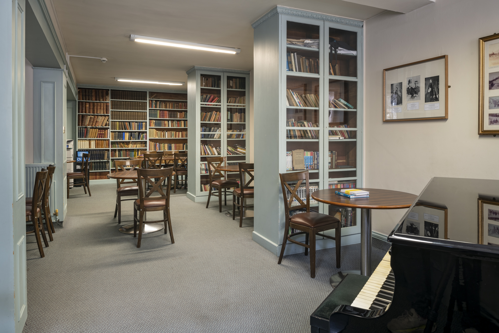 A room with several large bookshelves and book cases, and many circular tables and chairs. A piano is in the foreground.