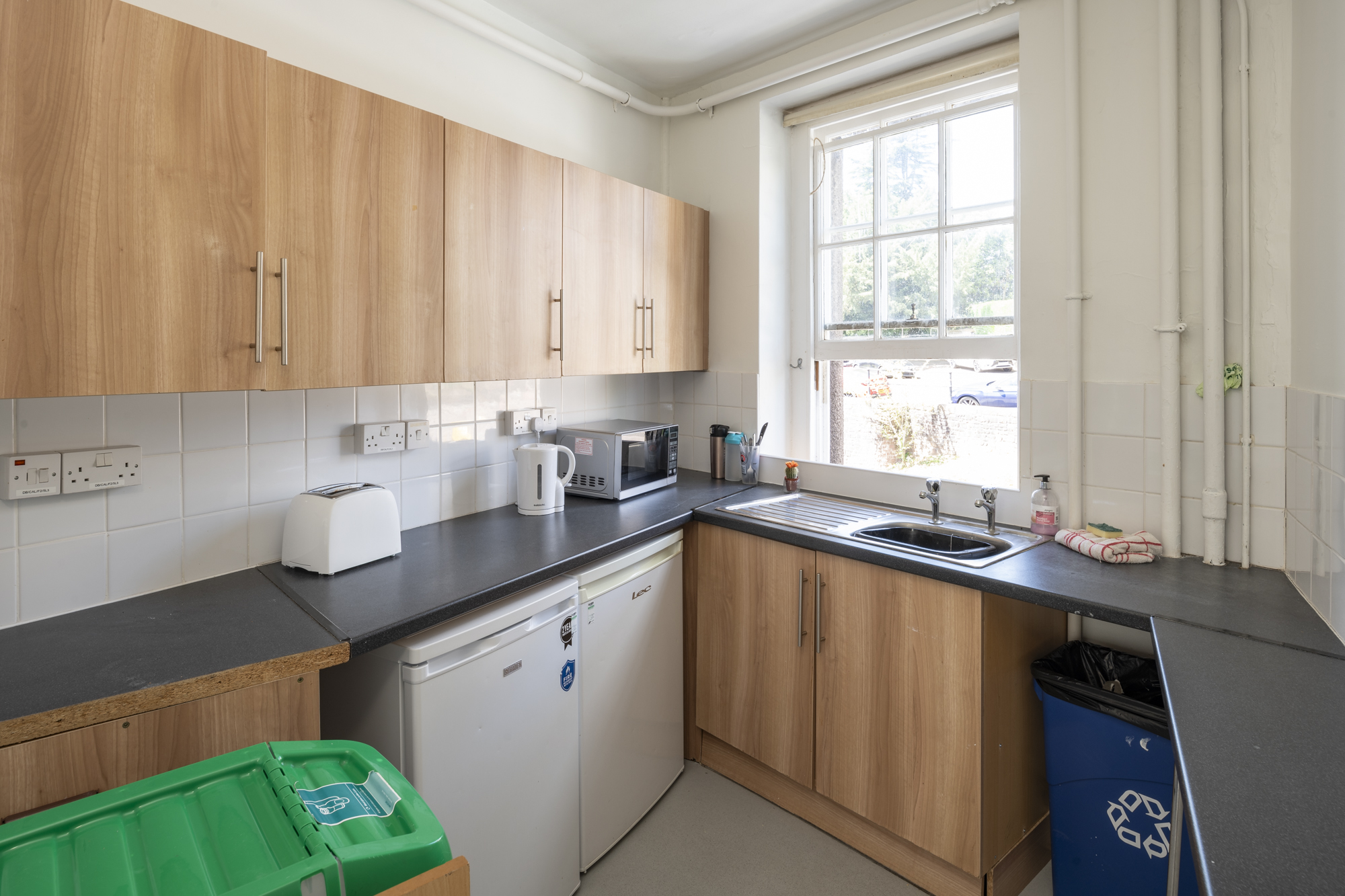 A kitchen with two fridges, recycling bins, a toaster, a microwave, a kettle, and cupboards.