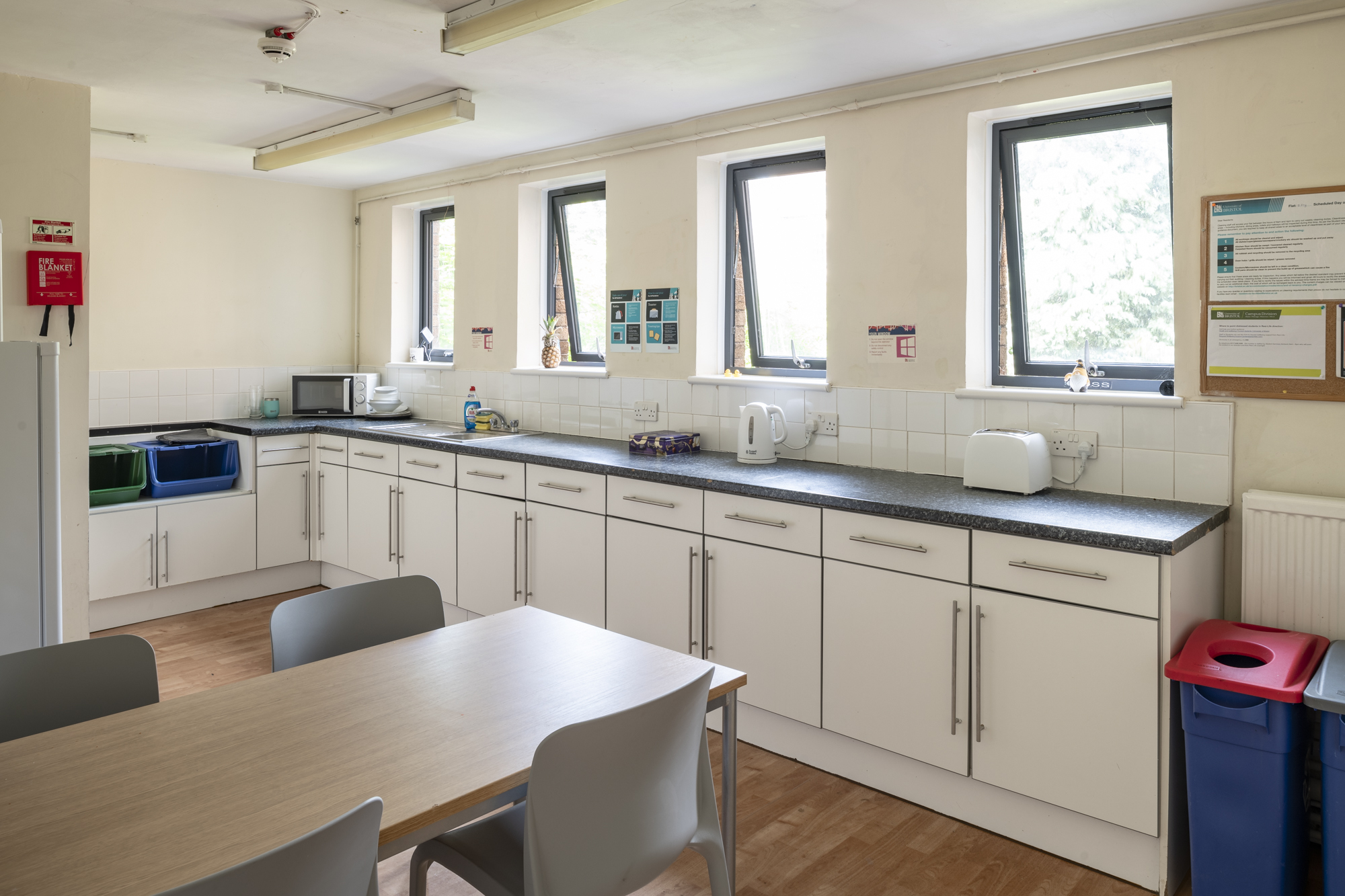 A room with a table with three chairs around it, two fridges, a fire blanket, a sink and lots of cupboards and drawers.