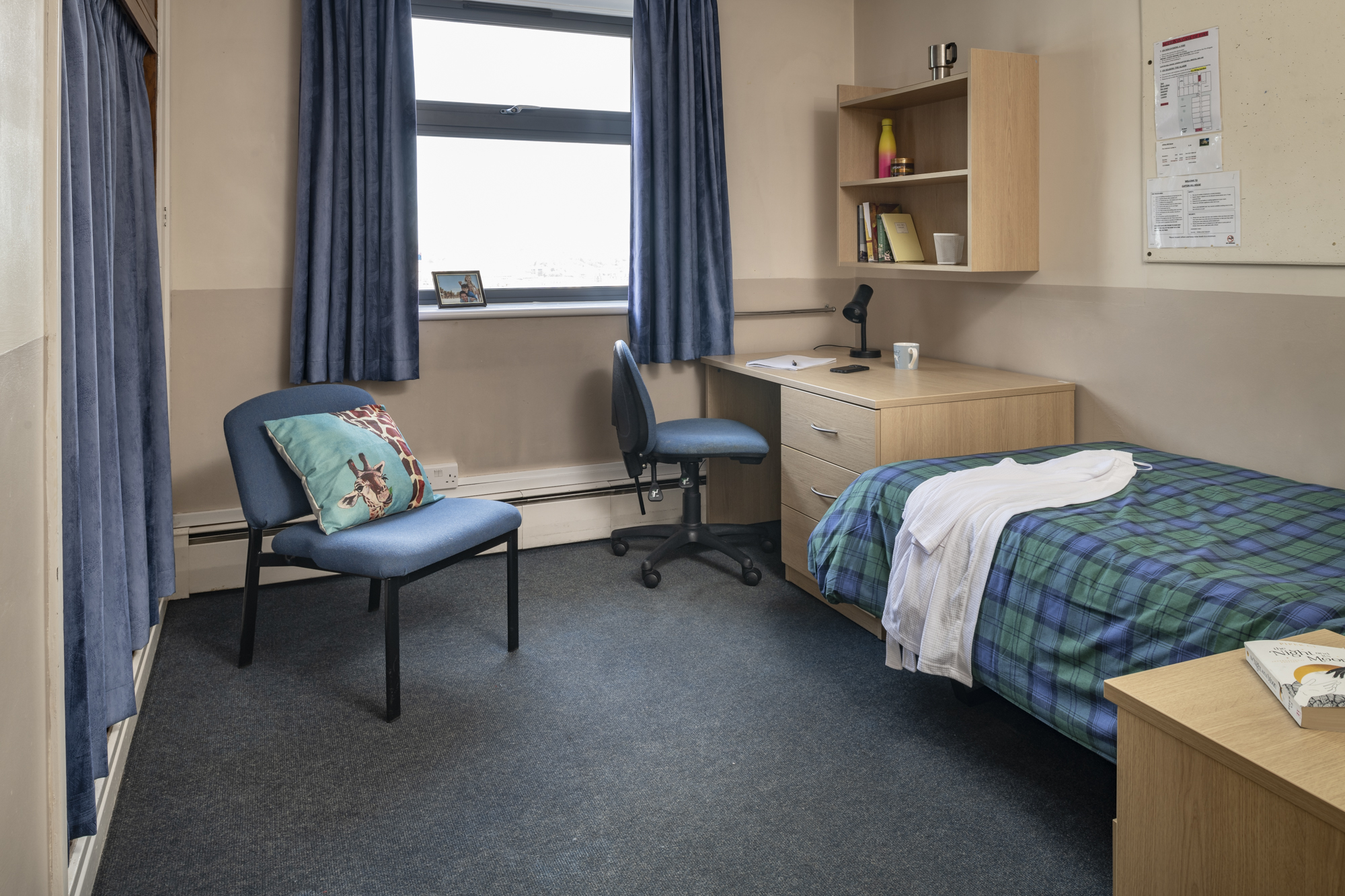 A room with a desk and office chair in one corner and a bed opposite it. There is a set of shelves over the desk, and a cupboard covered by curtains on the opposite wall with a chair nearby.