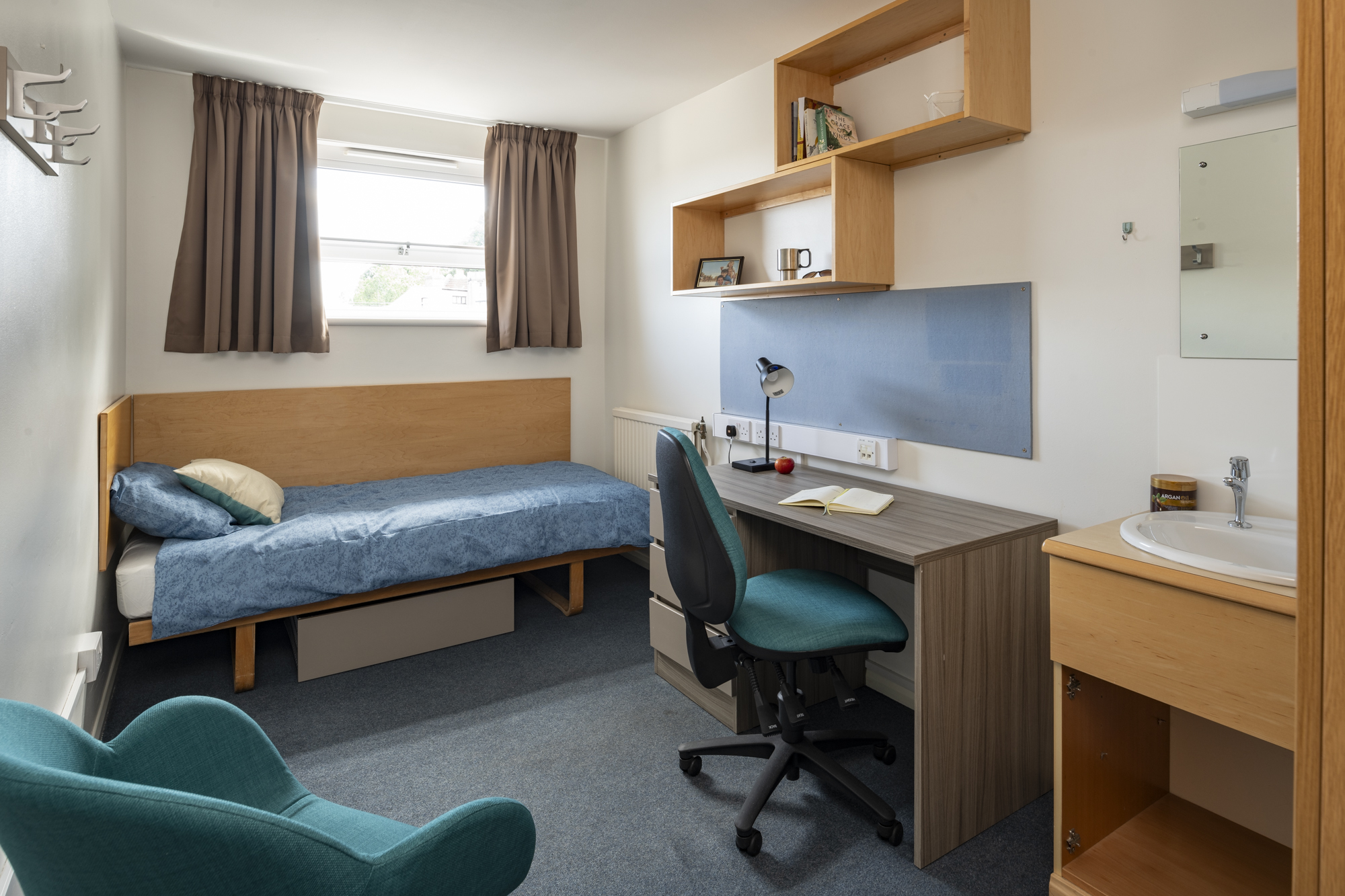 A room with a single bed along the fart wall and a window above. Along the right hand wall is a desk with deskchair and wall mounted shelves above. Next to this is a unit with a sink and a mirror above. On the left of the room is an armchair.