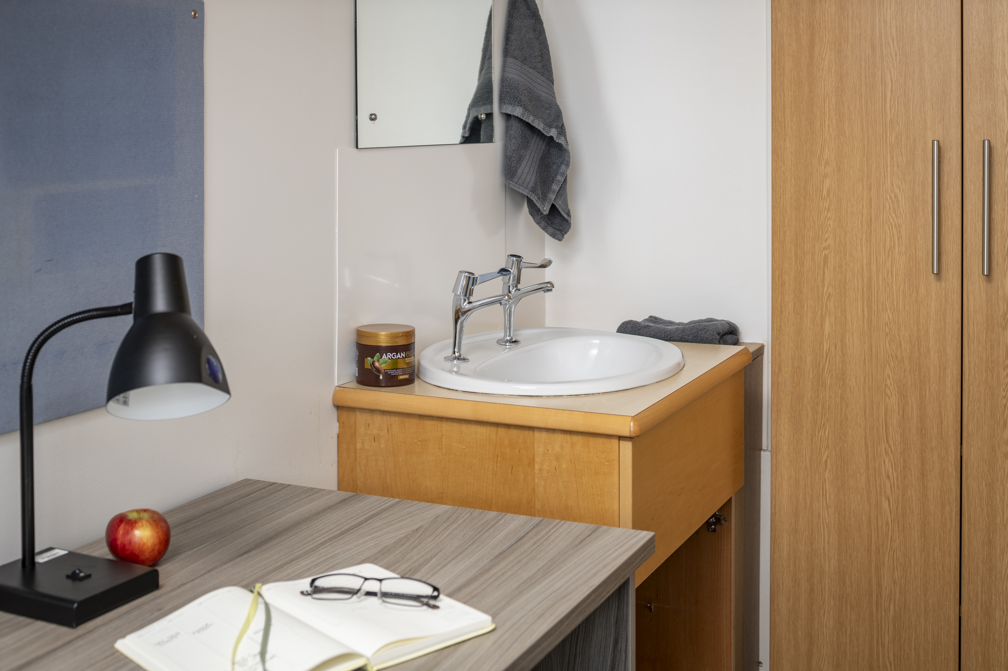 A desk with an artfully placed book upon which a pair of glasses are placed. behind this is an apple. Behind the desk is a unit with a two-tap sink and a mirror above. Next to this, along the back wall, is a wardrobe.