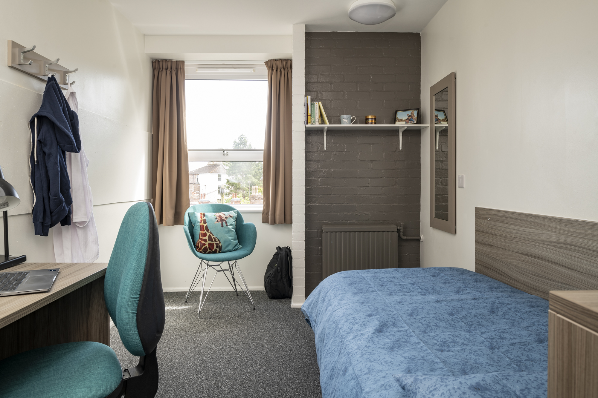 A room with a window at the far end and a legged armchair in front of it. On the left of the room is a desk with arm chair, behind this are wall mounted coat pegs. On the right, against the wall is a single bed. Behind this is a wall mounted shelf and mirror.