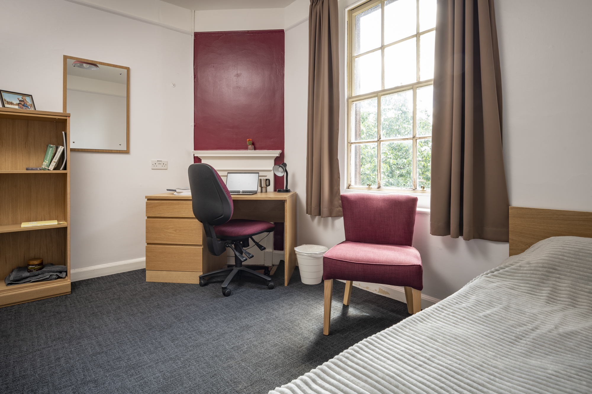 A bed in one corner and a desk with desk chair nin the opposite corner. Between the desk and chair is a window with a chair in front of it. To the left of the desk is a mirror and a set of shelves.