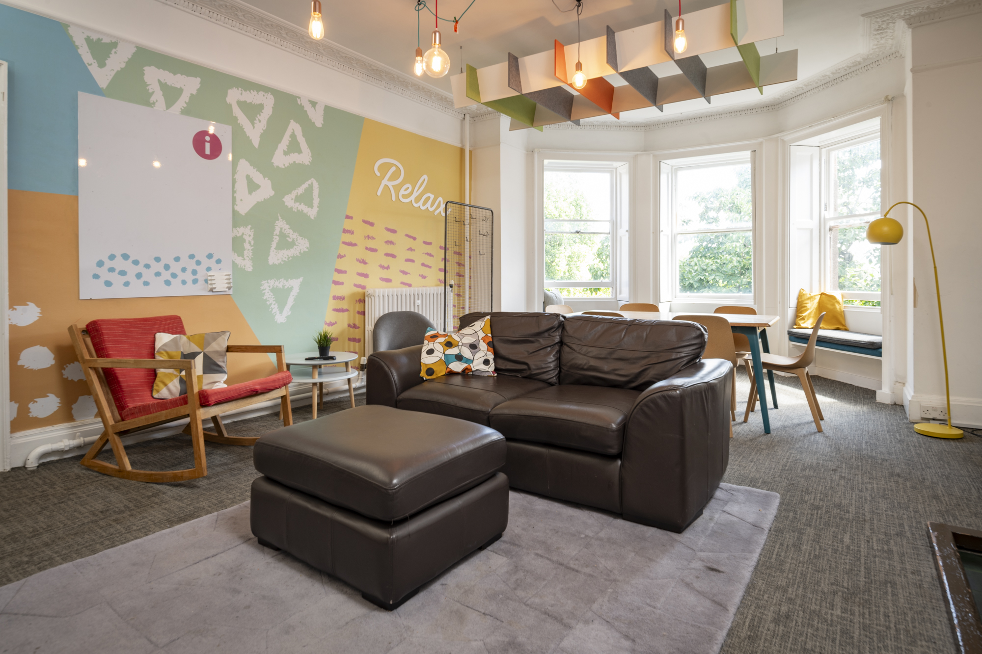 A sunny room with colourful walls. In the centre is an armchair and footrest. At the back by the windows is a table and chairs. There are some armchairs along the wall.