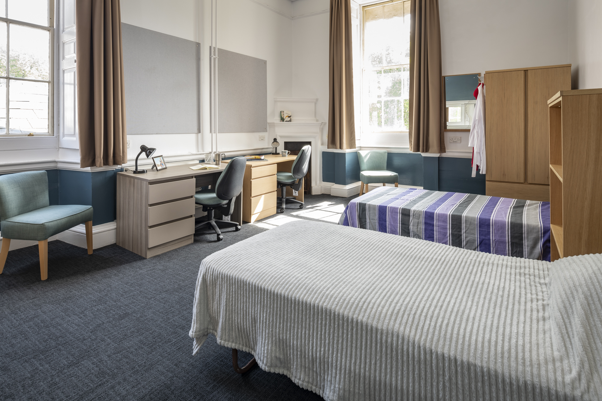 A room with two single beds, two desks with office chairs, an armchair and a cupboard.