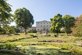 A large four-storey sandstone buidling with a large garden in front of it. The garden has several trees and a pond with a bench nearby.