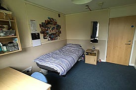 A room with a single bed, a desk with an office chair, a mirror on a wall by the bed and a set of shelves over the desk.