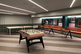 A room with a table football table, a ping pong table, and several tables with stools and chairs around them.