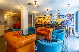 Two sofas and four armchairs around a coffee table, with a long high table with 18 stools around it behind them. The wall behind that has the word 'hive' written on it.