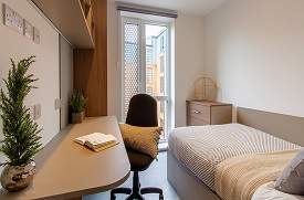 A room with a double bed in one corner and a desk with an office chair opposite. There are shelves above the desk and a small chest of drawers at the foot of the bed.