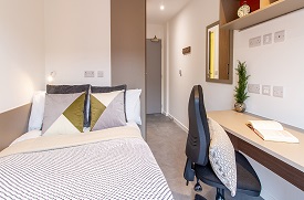 A room with a double bed in one corner and a desk with an office chair opposite. There is a cupboard behind the bed and a mirror on the wall opposite.