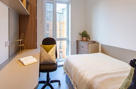 A room with a double bed in one corner and a desk with an office chair opposite. There are shelves above the desk and to its side.