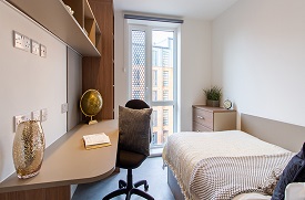 A room with a double bed in one corner and a desk with an office chair opposite. There are shelves above the desk and to its side.
