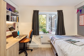 A room with a double bed in one corner and a desk opposite. The desk has an office chair next to it and shelves above it.