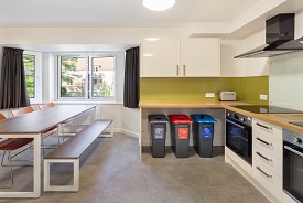 A kitchen with two ovens and hobs, three recycling bins, and a table with three chairs and a bench around it.