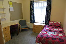 A room with a single bed in one corner and a small chest of drawers next to it. Against the opposite wall there is an armchair and a desk with an office chair.
