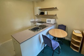A kitchen with a sink visible and shelves. A table with two chairs to the right of that.