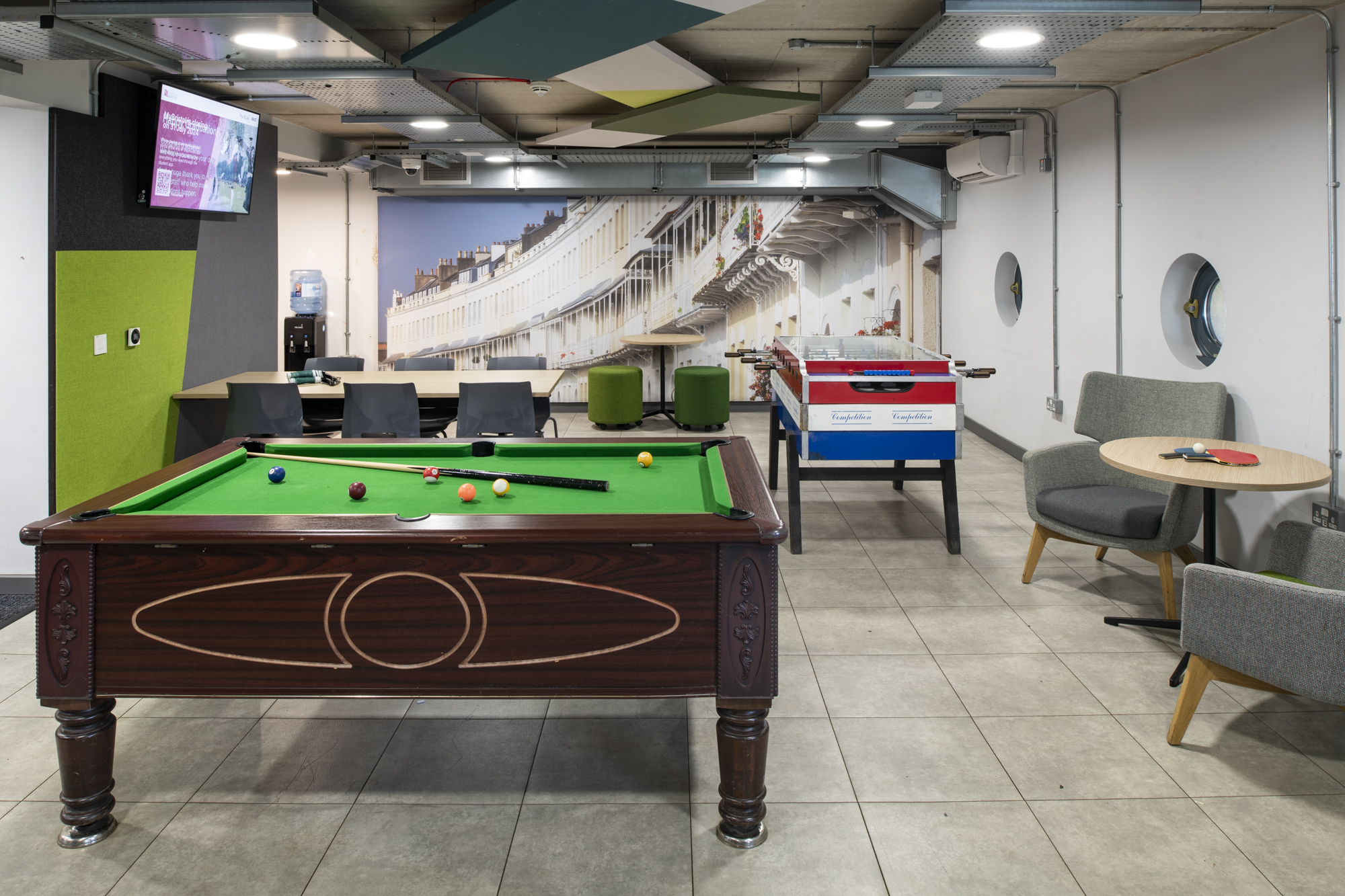 A room a snooker table, and table football. Tables and chairs and a televison mounted high on one wall.