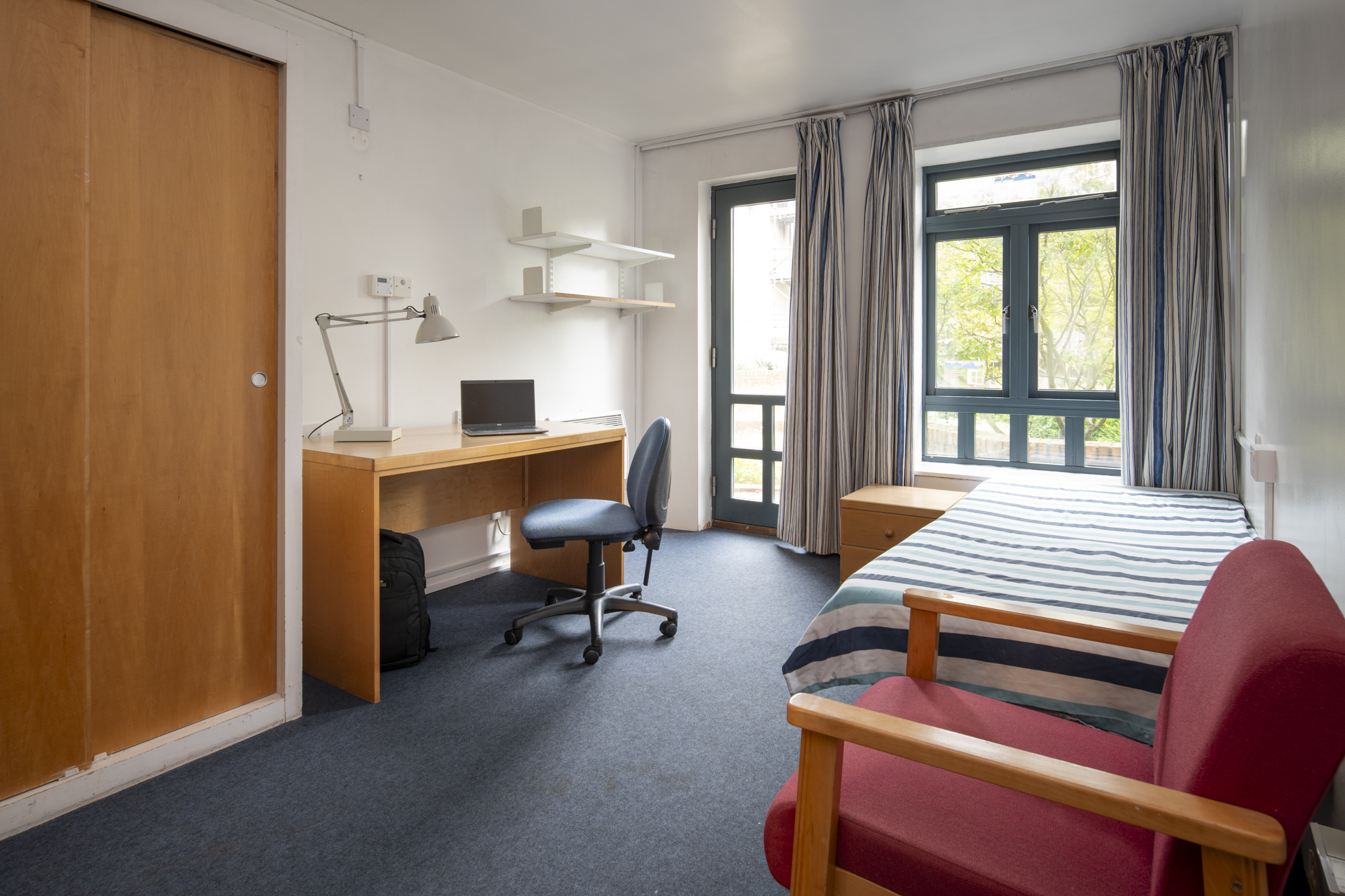 In the centre of the image is a storage unit with shelves, a sink and sideboard and a small two tier chest of draws beneath. To the right of the unit is an inbuilt wardrobe. To the left is the open door of the ensuite bathroom.