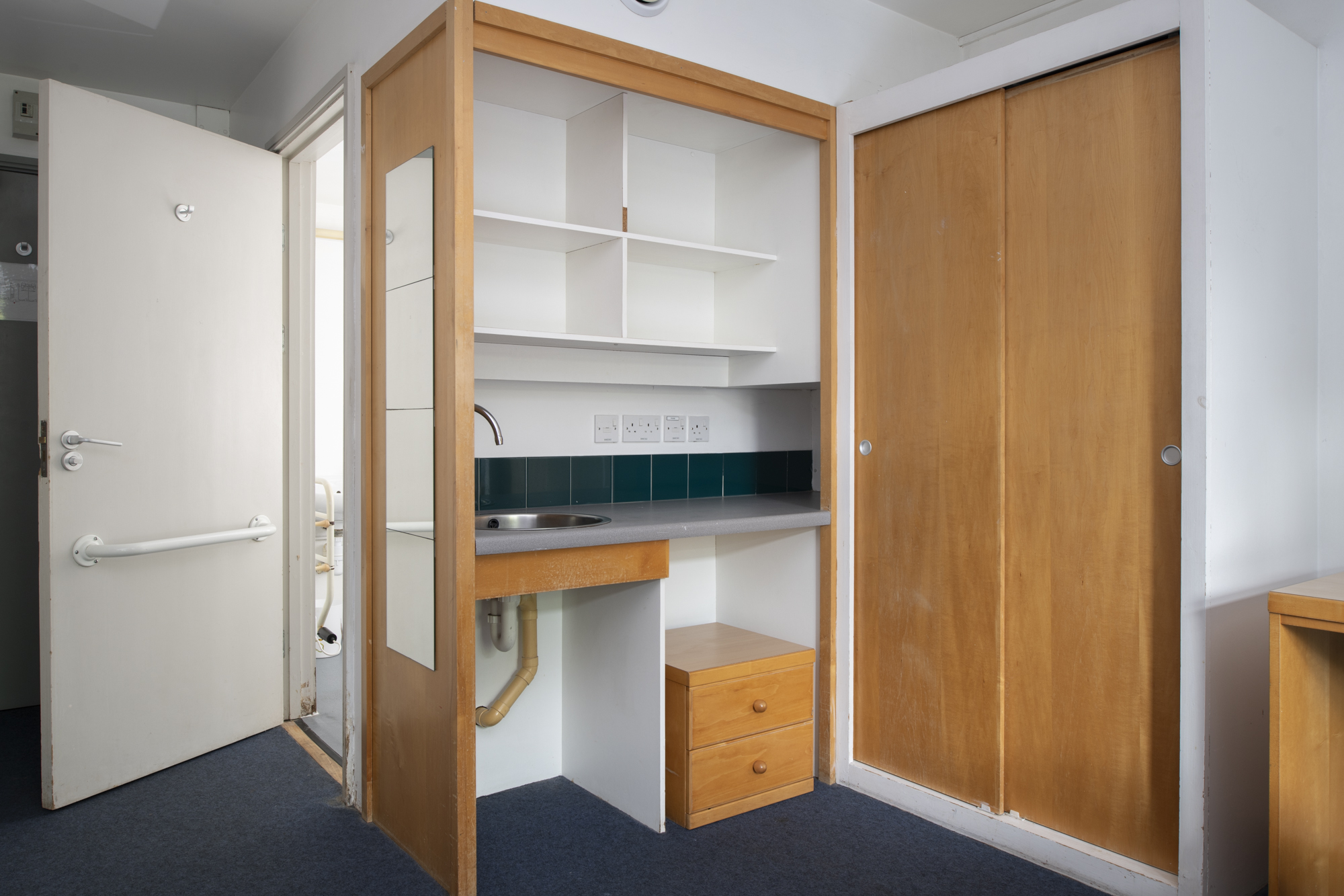 A room with a single bed against one wall, with an armchair at the foot of the bed, and a desk, desk chair and shelves against the opposite wall. Inbuilt storage is to the left of the desk.