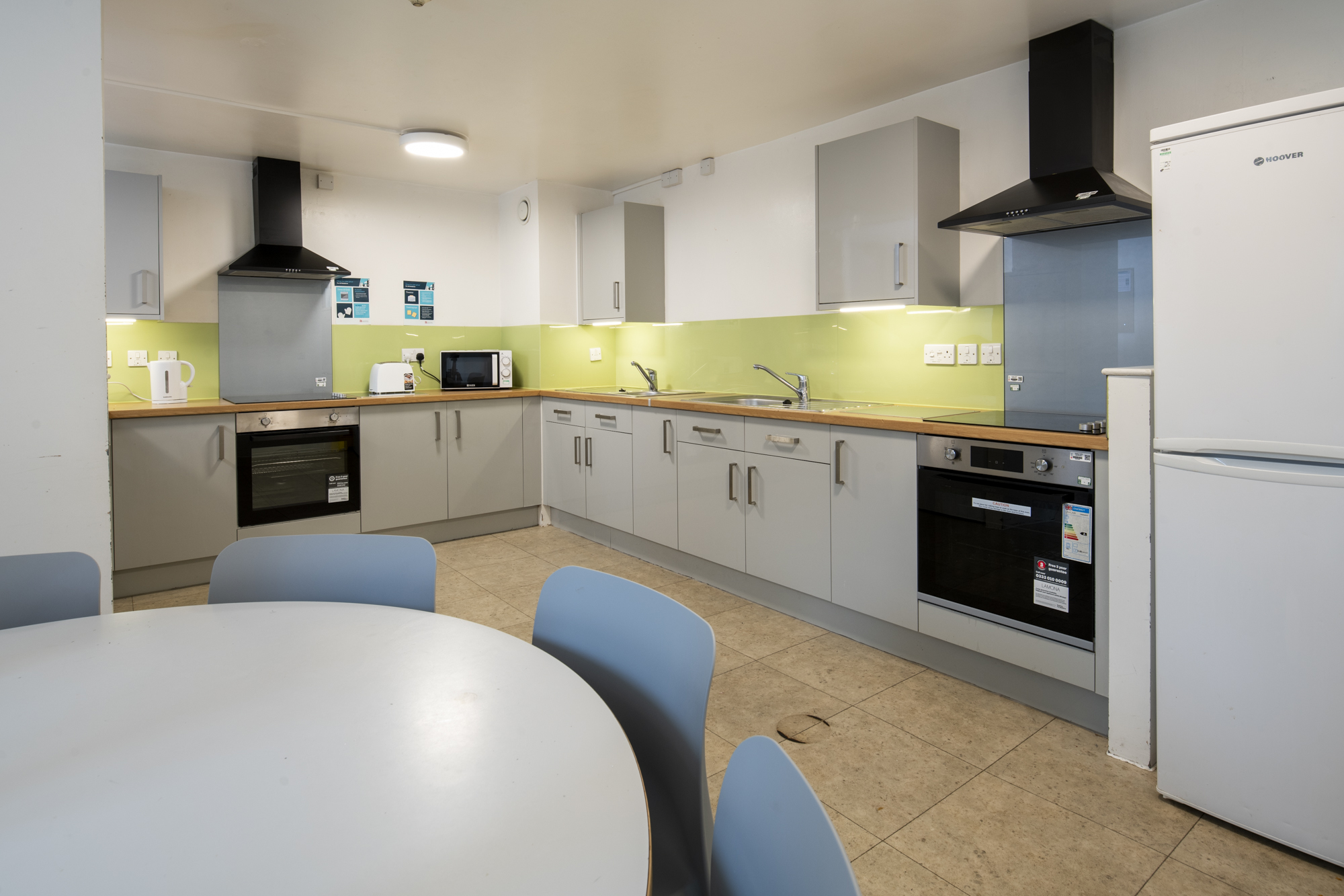 A kitchen with half of a circular table in the foreground. Behind are units with ovens, hobs, sinks, a fridge/frezer, and lots of cupboards.