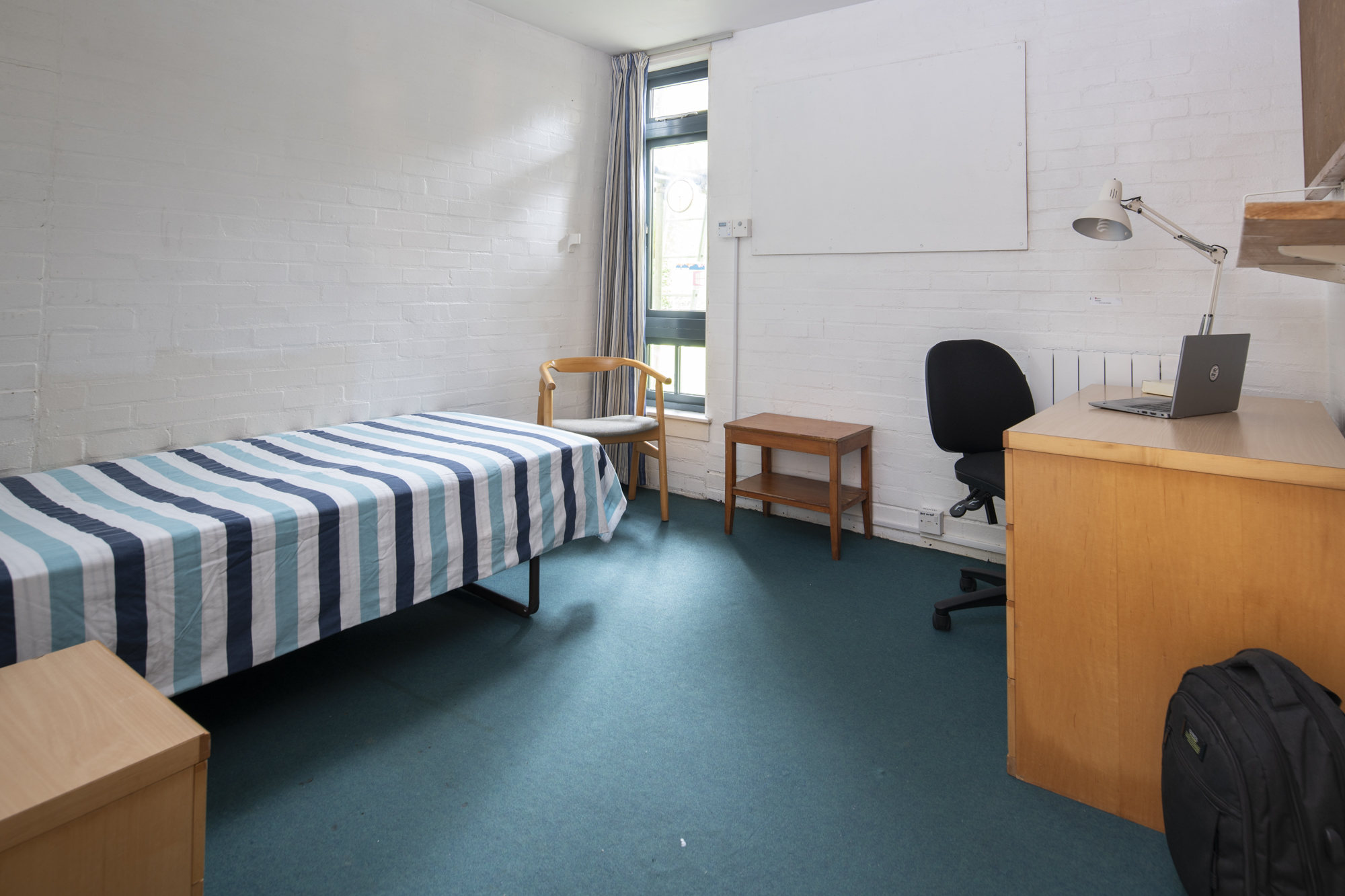 A room with a single bed against one wall and a bedside table beside it. On the opposite wall is a desk with desk chair and shelves above.