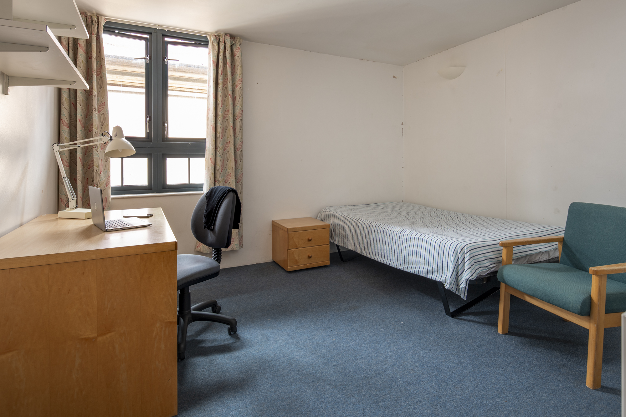 A room with a single bed in one corner and a desk opposite. There an armchair at the foot of the bed, an office chair next to the desk and a shelf above it.