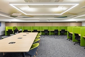 A room with a large desk with ten armchairs around it, and lots of smaller desks around the edge of the room. Each desk has an office chair and is separated from the others with a divider.