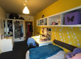 A room with a double bed in one corner and a desk at the foot of the bed. There is an office chair next to the desk and a shelf along the wall over the desk and bed. In the opposite corner there is a wardrobe and a chest of drawers.