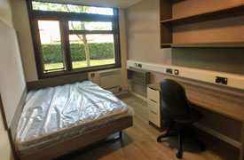 A room with a double bed in one corner and a desk against the opposite wall. The desk has an office chair next to it and a pair of shelves over it.