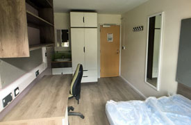 A room with a desk against the left wall, with a chair nearby and a set of shelves over the desk. On the opposite side of the room there is a bed and a mirror on the wall at the foot of the bed. At the far end of the room there is a wardrobe next to a sink which has another mirror over it.