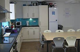 A kitchen with an oven and hob, a fridge freezer and lots of cupboards. There is a dining table nearby with six chairs around it.