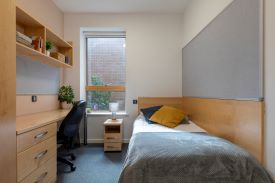 A room with a single bed in one corner, and a desk against the opposite wall. The desk as an office chair nearby, a set of shelves anove it and a set of drawers beneath it.