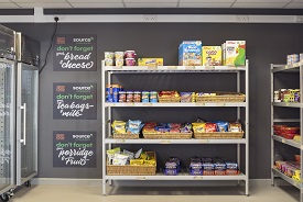 Shelves with food displayed, including breakfast cerals, biscuits, bread, crisps and sweets. Signs on the wall next to them say 'Don't forget your bread and cheese', '...your teabags and milk', and '...your porridge and fruit'.