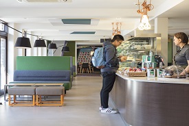 A food shop counter with a woman stood behind the till and a man stood on the other side. Behind him there are sofas, coffee tables and stools.